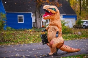 Person in inflatable dinosaur costume walking along suburban street with a Halloween candy bag. High quality photo